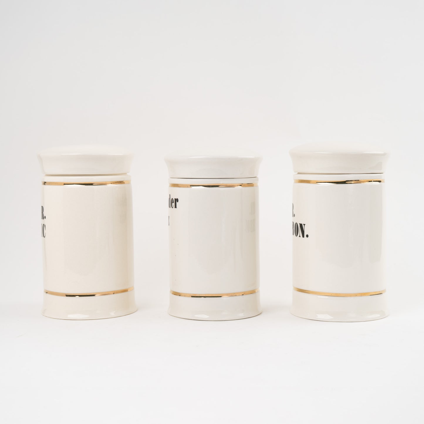 Set of three off-white ceramic apothecary jars with gold stripes and black vintage lettering with matching lids 