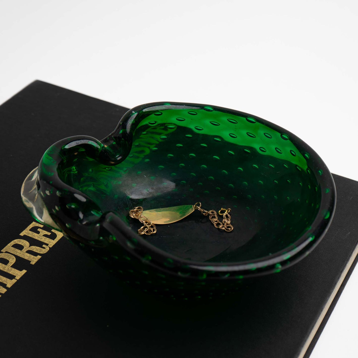 A dark emerald green, murano glass bowl catchall resting on a black book with gold lettering. Inside the bowl is a gold bracelet.